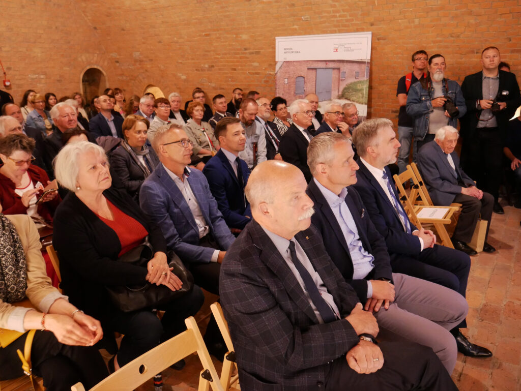 Liczna grupa słuchaczy siedzi na krzesłach, słuchając prowadzącego. W tle plakat ze zdjęciem wejścia do jednej z dawnych cel.