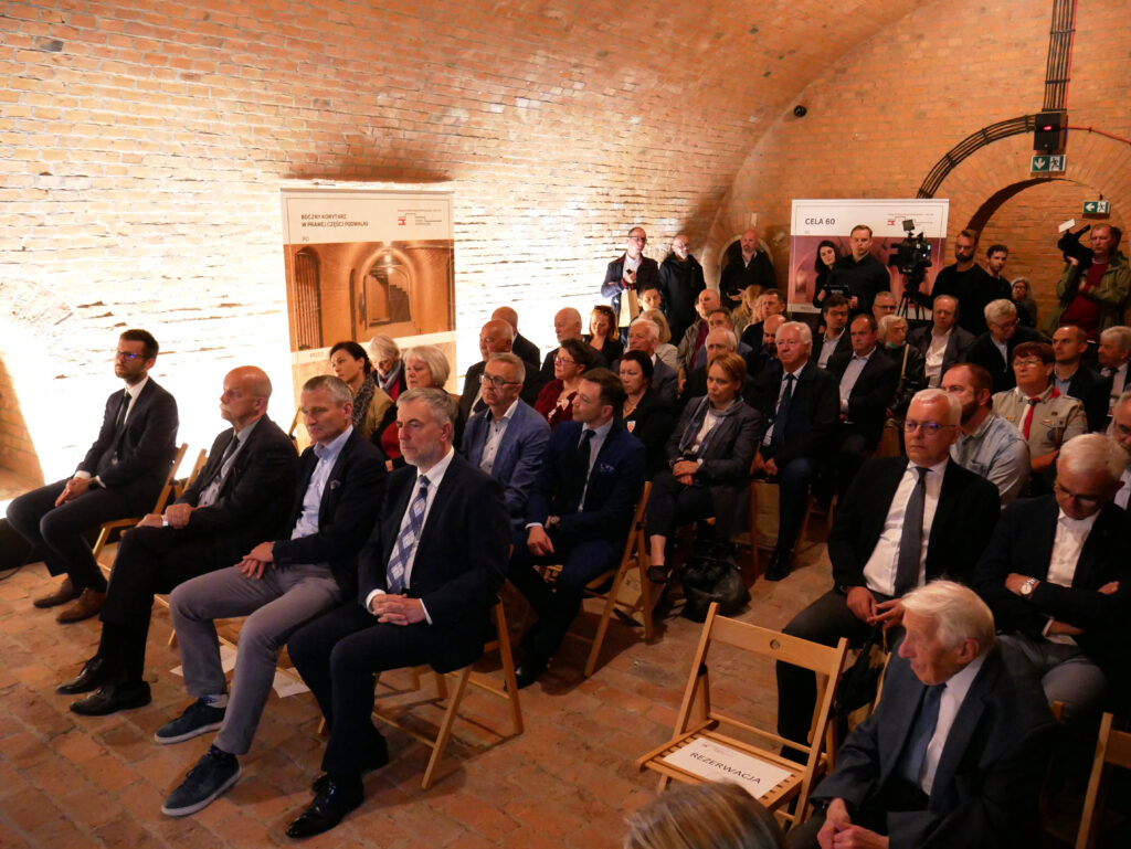 Liczna grupa słuchaczy siedzi w dawnej celi. W tle plakaty.
