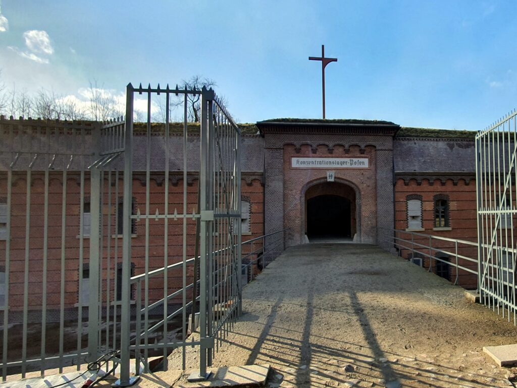 Na pierwszym planie metalowy płot i otwarta brama. W tle wejście na teren Fortu VII.
